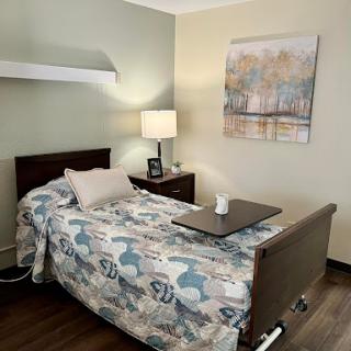 A comfortable hospital or patient room featuring a bed with a decorative blanket, a bedside lamp, and wall art.