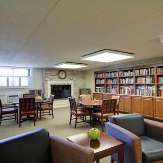 A cozy library reading room with shelves of books, tables, and chairs arranged around a fireplace.