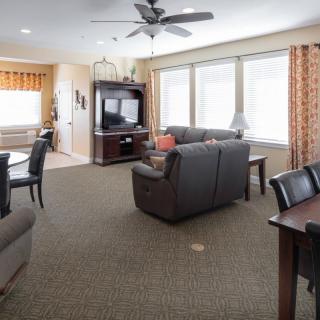 A cozy living room with a sofa set, dining table, and entertainment unit, featuring large windows and a ceiling fan.