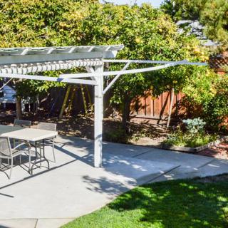 A sunny backyard featuring a pergola over a patio with outdoor furniture, surrounded by lush garden greenery.