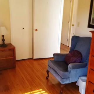 A cozy living room featuring a blue armchair and a wooden nightstand with a lamp, set against a backdrop of wooden floors and a white closet.