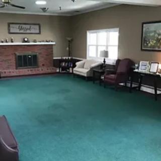 A spacious living room with a green carpet, featuring a fireplace, chairs, a sofa, and decorative artwork on the walls.