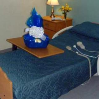 A hospital room featuring a bed with medical equipment, a gift basket on the table, and a lamp on the nightstand.