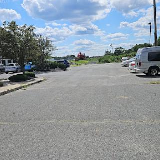 A spacious parking lot under a partly cloudy sky, featuring several parked vehicles and greenery in the distance.