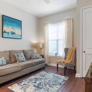 A cozy living room featuring a sofa with decorative pillows, an armchair, and a rug. A lamp stands near the window, which is adorned with light curtains, beneath a piece of wall art.