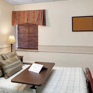 A tidy hospital room with a bed, adjustable side table, lamp, and window blinds.