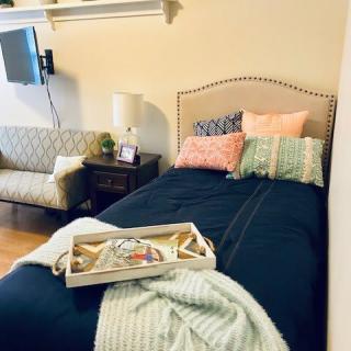 A cozy bedroom with a neatly made bed adorned with colorful pillows and a blanket. A tray is placed on the bed, and there's a lamp on the nightstand next to a small couch.