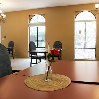 A cozy dining area featuring a table with a flower vase, surrounded by chairs and illuminated by natural light from large windows.