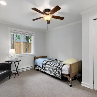 A minimalist bedroom featuring a single bed, ceiling fan, armchair, and a window providing natural light.