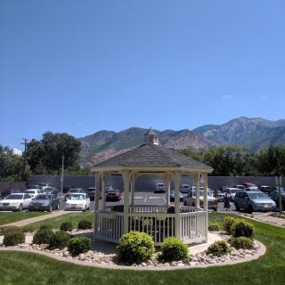 A scenic view of a park with a gazebo, surrounded by parked cars and mountains under a clear blue sky.