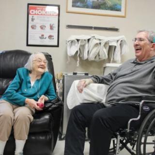 Two elderly individuals sharing a light-hearted moment indoors, with one seated in a wheelchair.