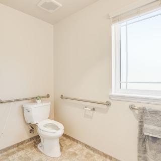 A bright, minimalist bathroom featuring a toilet, grab bars, and a window.