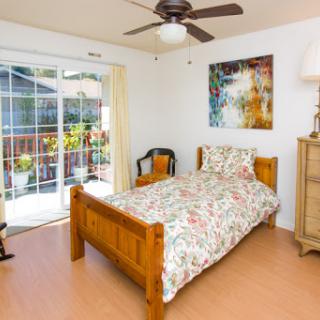 A cozy and well-lit bedroom featuring a wooden bed with floral bedding, complemented by a fan and artwork on the wall.