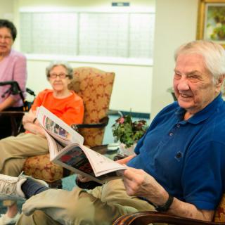 A group of seniors enjoying a moment together, with one man laughing while reading a newspaper.