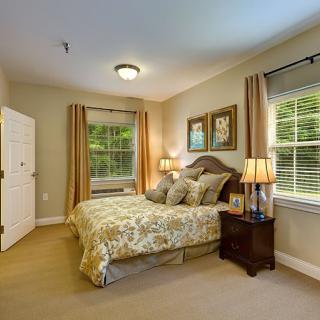 A cozy bedroom featuring a large bed with decorative pillows, bedside tables with lamps, and wall art. The room has carpeted flooring and is well-lit by natural light through the windows.