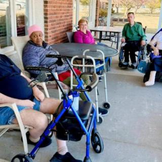 A group of elderly individuals seated outside a building, some using wheelchairs and walkers.