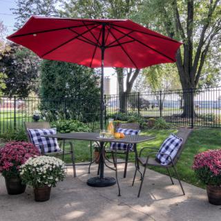 A cozy outdoor patio setup with a red umbrella, table, and chairs surrounded by potted flowers.