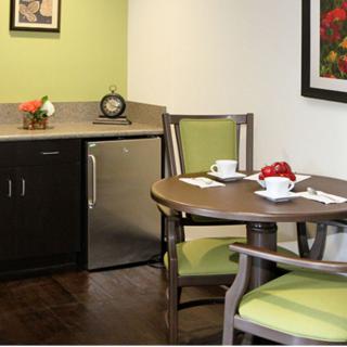 A small kitchenette area with a round table and chairs, a fridge, and decorative flower arrangements.