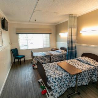 A hospital room with two beds, divided by a curtain, featuring overhead lighting and a window.