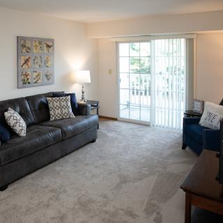A cozy living room with a dark leather sofa, decorative pillows, and a sliding glass door leading to a balcony.