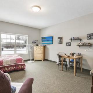A cozy bedroom with a large window, featuring a bed, dresser, table, and decorative shelves.