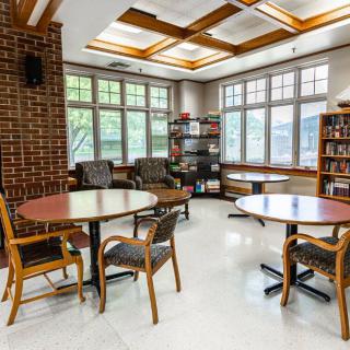 A cozy reading room with tables and chairs, surrounded by bookshelves and large windows.
