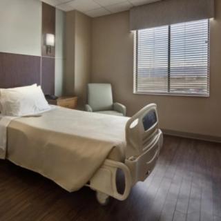 A modern hospital room featuring a single bed, a chair, and a window with blinds.