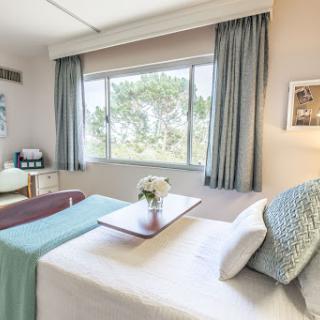 A well-lit hospital room featuring a comfortable bed, a small table with a flower vase, and large windows with curtains.