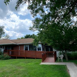 A cozy residential house with a wooden deck surrounded by greenery under a partly cloudy sky.