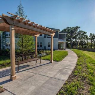 A peaceful outdoor area with a pergola-covered bench, a sidewalk, and a residential building in the background.