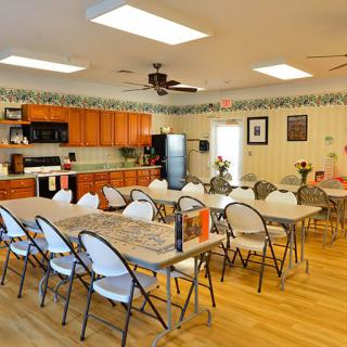 A spacious community meeting room with tables and chairs set up, adjacent to a kitchen with wooden cabinets.