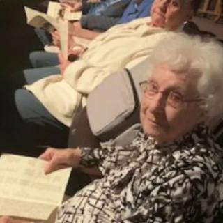 An elderly woman seated indoors, holding a book.