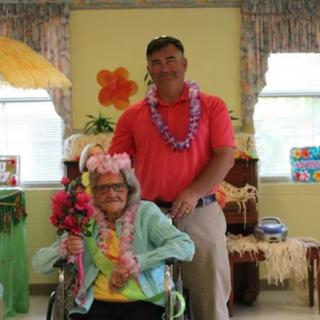 An elderly woman in a wheelchair and a man pose together, both wearing flower garlands in a room decorated with tropical themes.