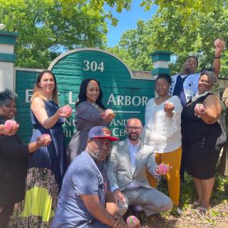 A group of people smiling and posing in front of a sign on a sunny day.
