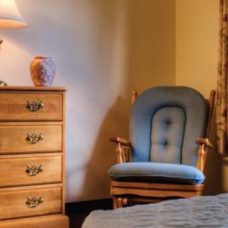 A cozy bedroom setting featuring a blue upholstered chair, wooden dresser, and a lamp providing soft lighting.