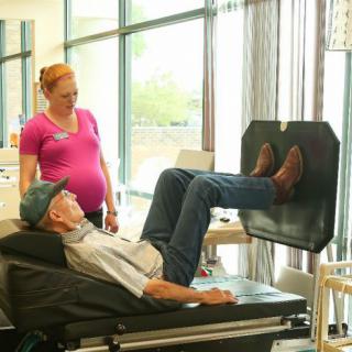 An elderly man performs a leg exercise with guidance from a healthcare professional in a rehabilitation setting.