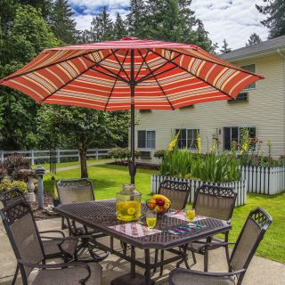 A cozy patio setting with a dining table and chairs under a red-striped umbrella, surrounded by lush greenery.