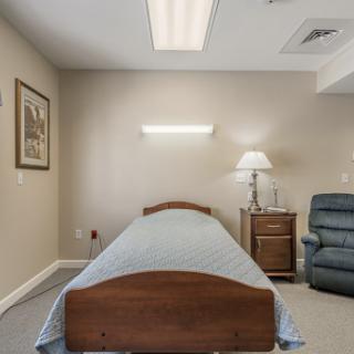 A simple and serene room featuring a single bed, a comfy chair, and soft lighting.