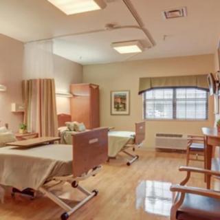 A well-lit hospital room with two patient beds separated by a curtain, featuring chairs and a window for natural light.
