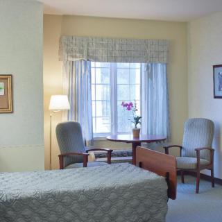 A cozy bedroom with a bed, chairs, and a table near a window, complemented by soft lighting from a floor lamp.