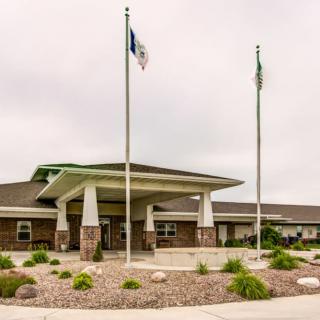A single-story building entrance with flagpoles and landscaped surroundings under a cloudy sky.