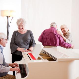 A group of elderly people enjoy music, with one playing the piano and two others dancing.