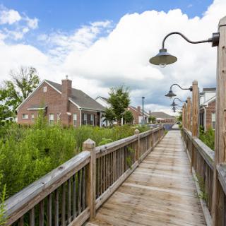 A scenic wooden pathway lined with houses, leading through lush greenery under a partly cloudy sky.