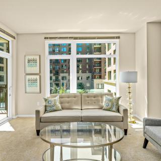 A modern living room with a stylish sofa and glass table, featuring a large window allowing ample natural light and offering an urban view.