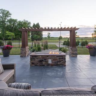 A beautifully designed patio with a fire pit and a pergola, surrounded by lush greenery at sunset.