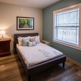 A cozy bedroom with a neatly made bed, nightstand, and window, featuring a soft lamp light and wall art.