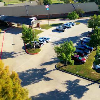 An aerial view of a parking lot filled with cars, framed by lush greenery and a building in the background.