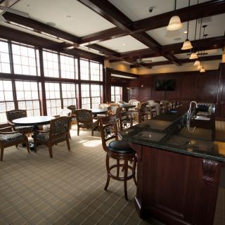 A cozy bar and lounge area with tables and chairs, featuring large windows and warm lighting.