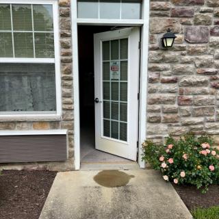 An open white door leading to the interior of a brick building, with flowers and a window nearby.