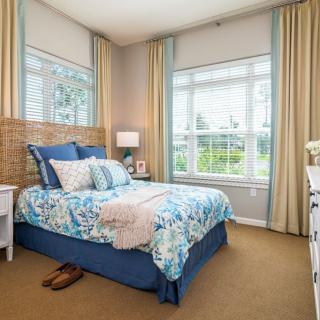 A cozy bedroom with large windows, featuring a bed adorned with blue and floral bedding and surrounded by light-colored curtains.